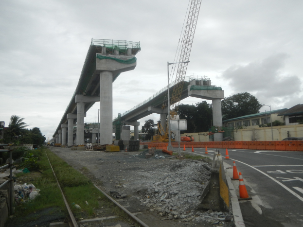 Baustelle mit einer Brücke im Hintergrund, Straße mit Verkehrskegeln markiert, Bahnschiene links, verstreute Steine und Gras, Bäume und Gebäude entlang der Straße und ein bewölkter Himmel.