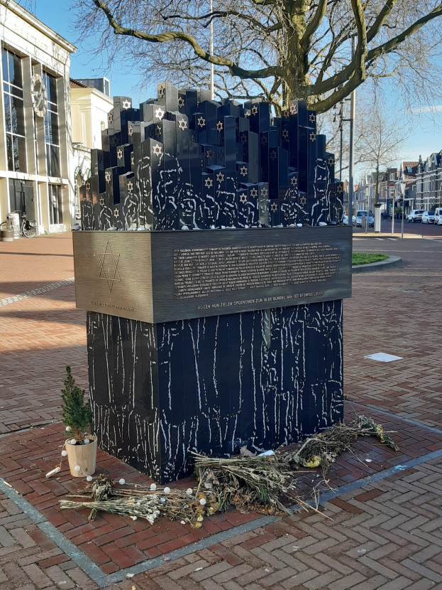 Denkmal in einem Stadtplatz fur Holocaust-Opfer, mit einem Baum davor, einer Topfpflanze auf dem Boden, verstreute trockene Blatter, umgeben von Gebauden, Fahrzeugen, Pfosten, Baumen und Himmel.