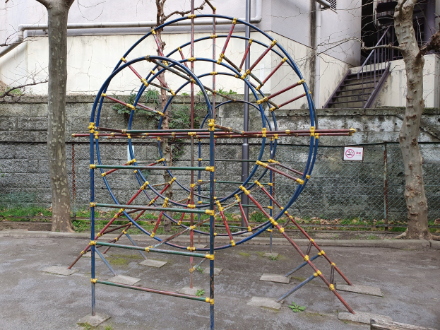 Spielplatz mit großem Metallklettergerüst, umgeben von Bäumen und einer Maschendrahtzaunmauer, mit Stufen mit Geländern, die zu einem Gebäude mit Fenstern und Rohren führen, einem Schild an einem Pfahl und grasbewachsenem Boden.