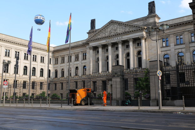 Großes modernes Gebäude mit vielen Fenstern, als Bundestag in Berlin, Deutschland identifiziert, umgeben von Straßeninfrastruktur, Fahrzeugen, Fußgängern, Flaggen und Bäumen unter einem bewölkten Himmel.