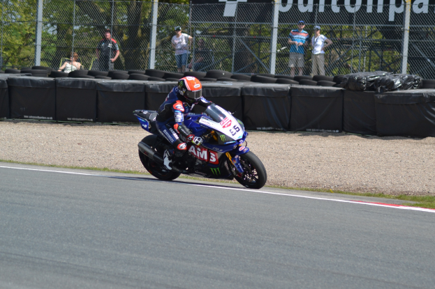 Ein Motorradfahrer in Helm und Handschuhen fährt auf einer von Gras, Reifen, Maschendrahtzaun und einem Banner gesäumten Rennstrecke, mit Bäumen und einem klaren blauen Himmel im Hintergrund.