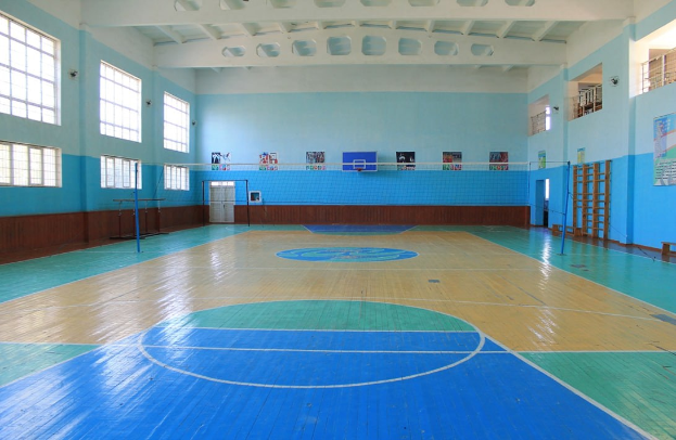 Großer Turnsaal mit blau-weißem Boden, Basketballfeld, Netz, Stützpfosten, einer Tür, Fenstern, Wandplakaten, einem Geländer und einer Dachkonstruktion.