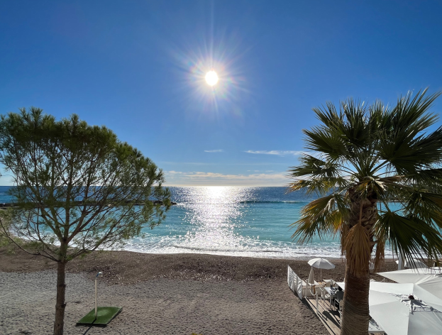 Ein Strand mit Palmen, Sonnenschirmen, üppiger Vegetation, einem strahlend blauen Himmel und einer scheinenden Sonne im Hintergrund, geeignet für eine Ferienwohnung an der französischen Riviera.