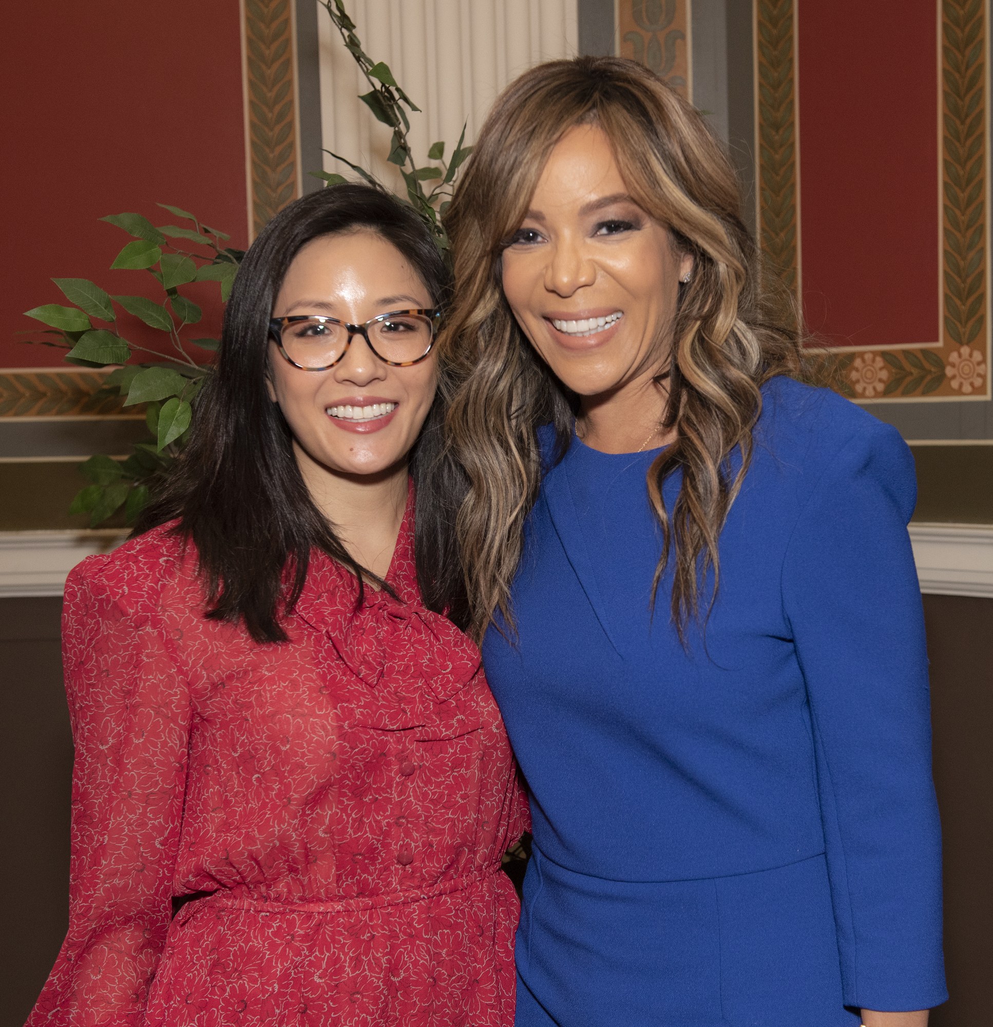 Zwei Frauen in roten und blauen Kleidern lächeln gemeinsam auf der 2019 Women in Film Pre-Oscar-Lunch-Veranstaltung, mit einer Pflanze und einem Vorhang im Hintergrund.