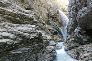 Kleiner Wasserfall, der an zerklüfteten Felsen in einem steinigen Tal hinabfließt, umgeben von grünen Hügeln unter strahlendem Sonnenschein.