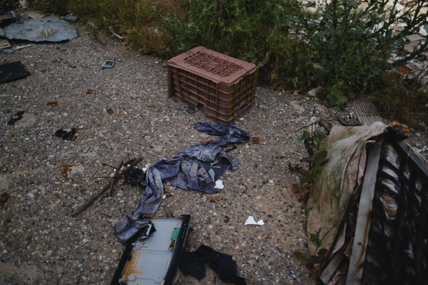 Ein Haufen Müll, darunter ein Plastikbehälter und Kleidung, liegt auf dem Boden neben einem Zaun auf einer Müllhalde, mit Pflanzen und Gras im Hintergrund.