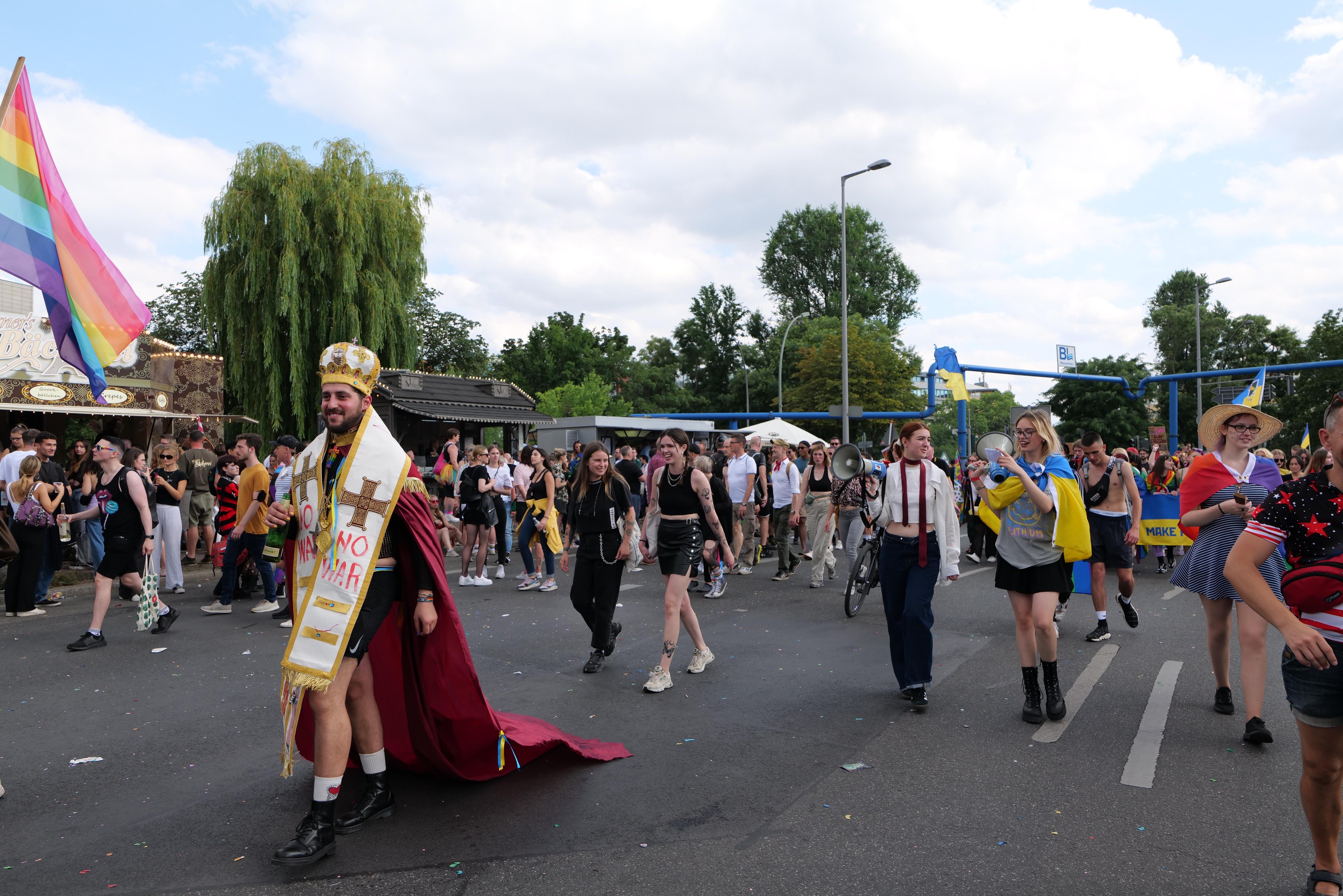 Eine Gruppe von Menschen marschiert auf der Pride Parade 2018 mit einer Regenbogenflagge und Musikinstrumenten, umgeben von Laternenmasten, Bäumen, Hütten und einem bewölkten Himmel.