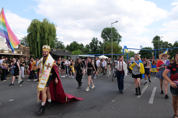 Eine Gruppe von Menschen marschiert auf der Pride Parade 2018 mit einer Regenbogenflagge und Musikinstrumenten, umgeben von Laternenmasten, Bäumen, Hütten und einem bewölkten Himmel.