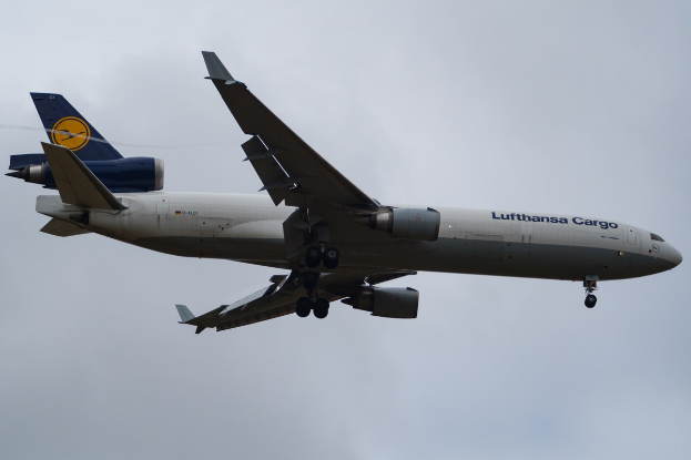 Ein Frachtflugzeug des Typs Boeing 777-300ER der Lufthansa Cargo im Flug, mit ausgefahrenen Tragflächen und aktivierten Triebwerken, mit sichtbarem Text auf dem Rumpf gegen einen wolkengefüllten Himmel.