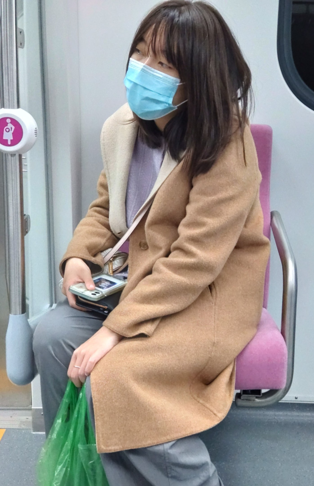 Eine Frau mit Gesichtsmaske sitzt in einem Zug und hält ein Mobiltelefon und eine Tasche in der Hand, wobei eine Tür auf ihrer linken Seite zu sehen ist.