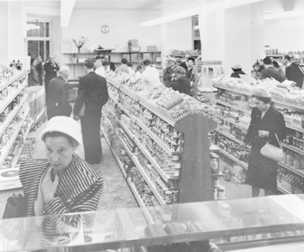 Schwarze und weiße Fotografie von Menschen beim Einkaufen in einem Supermarkt mit Regalen voller Lebensmittel, einige halten Taschen, eine Uhr an der Wand und Deckenbeleuchtung.