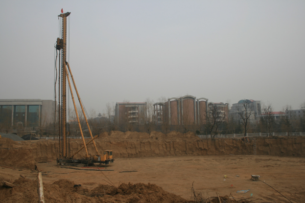 Eine Baustelle mit einem Kran in der Mitte, umgeben von Gebäuden, Bäumen und einem klaren blauen Himmel, mit Erde bedecktem Boden und verstreuten Gegenständen.