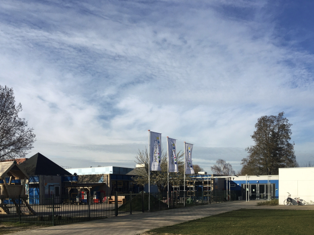 Großes Gebäude mit einem grasigen Bereich davor, umgeben von einem Metallzaun, mit einem Weg, Bäumen, Pfählen, Bannern, einem geparkten Fahrrad, näheren Häusern und einem bewölktem Himmel, als das neue Kinderspielgelände an der Schule identifiziert.