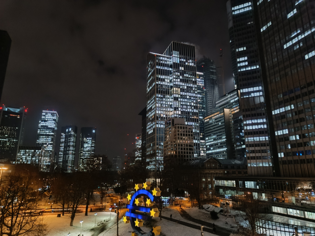 Hauptsitz der Europäischen Zentralbank in Frankfurt, Deutschland bei Nacht, mit beleuchteten Gebäuden, Bäumen, Straßenlaternen, Lichtern und Fußgängern auf der Straße unter einem bewölkten Himmel.