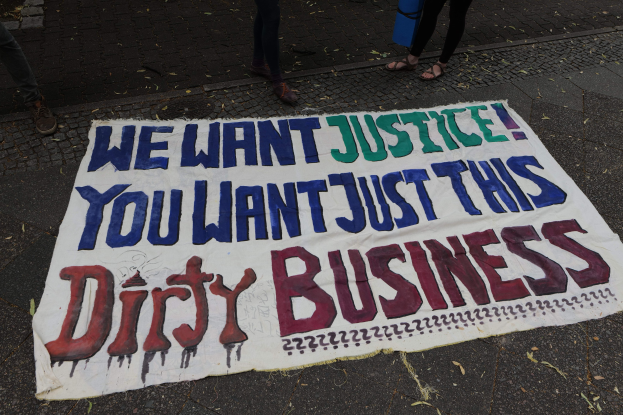Banner auf einer Straße mit dem Text "Wir wollen Gerechtigkeit, du willst nur diesen schmutzigen Deal" und zwei Personen oben stehend, eine hält ein blaues Objekt.