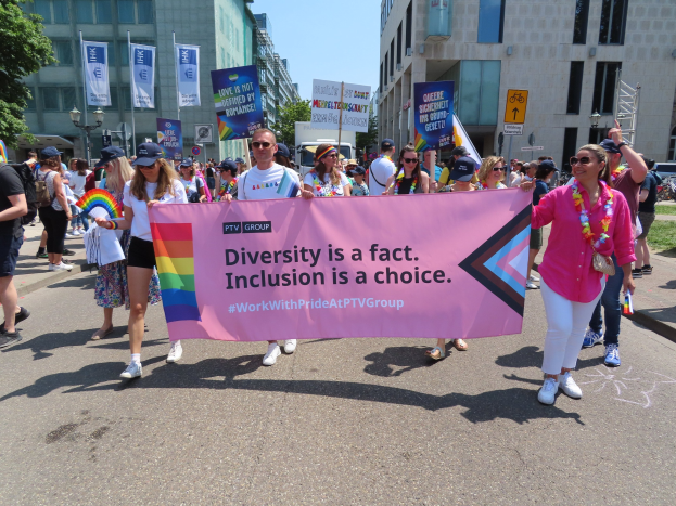 Eine Gruppe von Menschen marschiert auf einer Straße, hält ein Banner mit der Aufschrift "Diversität ist eine Tatsache, Inklusion ist eine Wahl" in den Händen, einige tragen Mützen und Schutzbrillen, andere halten zusätzliche Banner, vor einem Hintergrund aus Gebäuden, Bäumen, Laternenmasten und einem klaren blauen Himmel.