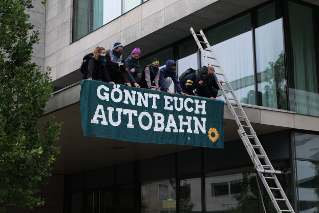 Gruppe von Menschen auf einem Gebäude, die ein Banner halten, mit einem Baum links und Glasfenstern im Hintergrund, die scheinbar für die deutsche Regierung protestieren.