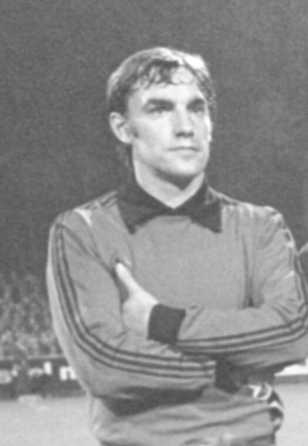 Schwarzes und weißes Foto eines Mannes auf einem Fußballfeld mit verschränkten Armen, umgeben von sitzenden Zuschauern, der als legendärer Torwart von Manchester United gilt, der im Alter von 89 Jahren verstorben ist.
