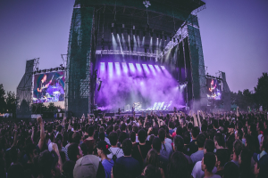 Eine große Menschenmenge auf einem Musikfestival schaut auf der Bühne mit Instrumenten und Bildschirmen, die vor einem Gebäude, Bäumen und einem klaren blauen Himmel aufgebaut sind.