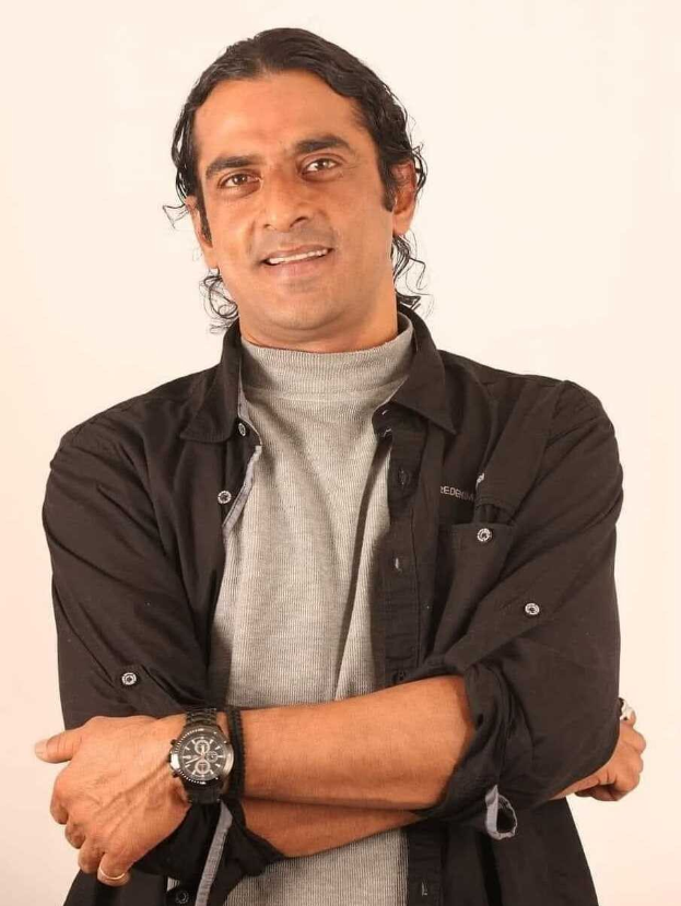 Ein Mann in einem schwarzen und aschfarbenen Kleid mit verschr├Ąnkten Armen steht vor einem wei├čen Hintergrund, tr├Ągt eine Uhr und sieht nachdenklich aus.