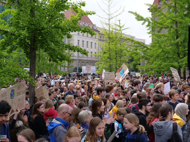 Große Menschenmenge protestiert vor einem Gebäude in Berlin, hält Schilder hoch, mit Bäumen, Fahrzeugen, einem Lautsprecher und Himmel.