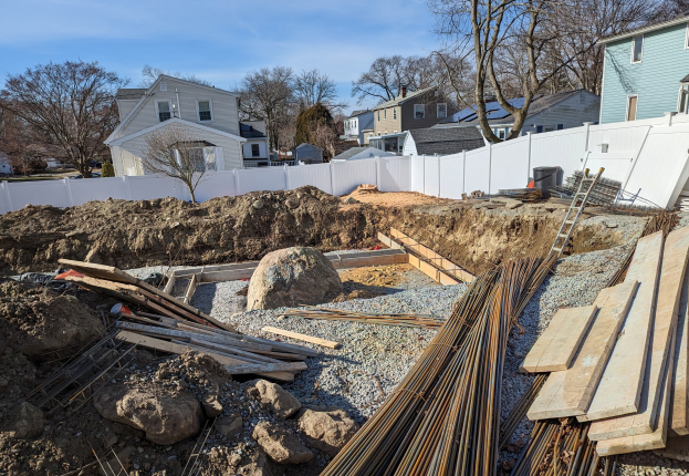 Baustelle mit Holzplanken, Steinen, Felsen, einer Leiter und einem weißen Zaun, mit Gebäuden, Bäumen, Fahrzeugen und bewölktem Himmel im Hintergrund.
