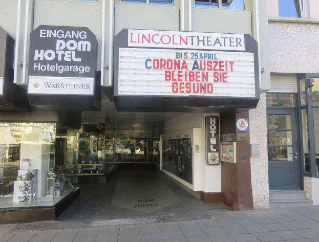 Außenansicht des Lincoln Theaters in Berlin, Deutschland, mit Glasfenstern und -türen und einer Tafel mit Text sowie einer belebten Stadtlandschaft im Inneren.