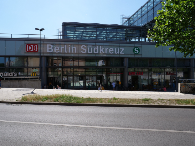 Großes gläsernes Berliner Südkreuz-Bahnhof mit Text an der Fassade, umgeben von Straßenlaternen, Fahrzeugen, Fußgängern, Fahrrädern, Bäumen und einem klaren blauen Himmel.