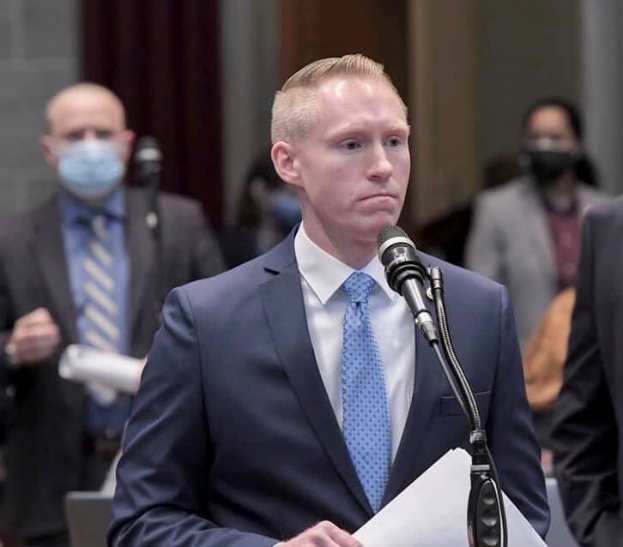 Mann in Anzug und Krawatte, der an einem Rednerpult spricht und Papiere in der Hand hält, vor einem Mikrofon und einer Wand mit Vorhang im Hintergrund.