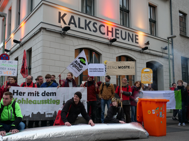 Eine Gruppe von Menschen mit Protestschildern vor einem Gebäude in Deutschland, mit zwei Personen im Vordergrund und einem Müllcontainer auf der rechten Seite.