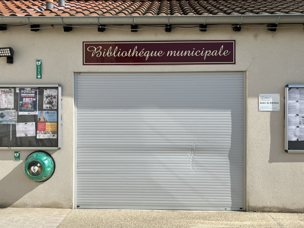 Ein Rathaus mit einer 'Bibliothèque municipale'-Schrifttafel, das eine mittlere Jalousie, eine Tafel mit Text, Plakate an der linken Wand und eine beleuchtete Beleuchtung zum Eingangspfad zeigt.