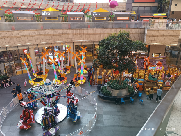 Ein Vergnügungspark in einem Einkaufszentrum mit verschiedenen Attraktionen, Bäumen, Menschen und Ständen, umgeben von Gebäuden oben und einem Wasserzeichen unten.