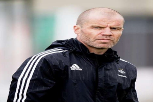 Ein kahlköpfiger Trainer in einer blauen Jacke mit weißen Streifen und weißem Kragen steht auf einem Fußballfeld und hat die Hände verschränkt und einen ernsten Gesichtsausdruck.