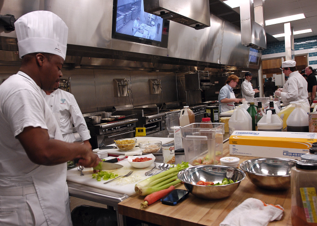 Eine Gruppe von Köchen in einer Küche, die auf einem Schneidbrett Essen zubereiten, wobei ein Koch im Vordergrund eine weiße Schürze und eine Mütze trägt und ein Messer hält und von Utensilien, Schüsseln, Gemüse, einem Mobiltelefon, Flaschen und anderen Gegenständen umgeben ist, mit einem Fernseher und Deckenleuchten im Hintergrund.