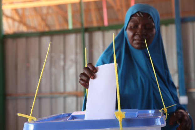 Eine Frau mit einem blauen Kopftuch steht und hält ein Papier in der Hand, vor sich eine Kiste mit Stöcken, vor einer Wand und einem Schuppen im Hintergrund.