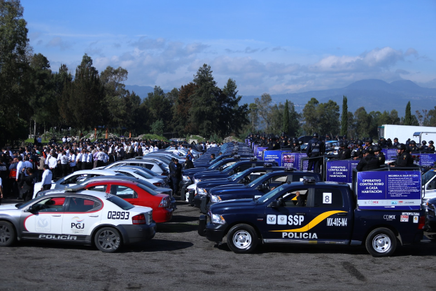 Große Anzahl von Polizeiwagen auf einem Parkplatz mit Menschen in der Nähe, Schilder mit Text im Vordergrund, Bäume und Hügel im Hintergrund unter einem bewölkten Himmel.