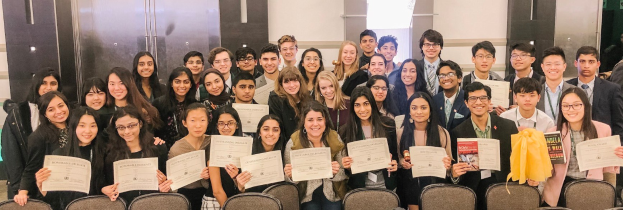 Gruppe von Menschen mit Urkunden, die zusammen in einem Raum mit Stühlen und einer Wand im Hintergrund stehen, wahrscheinlich feiern sie eine Abschlusszeremonie für das Studienjahr 2019-2020.