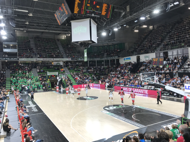 Basketballspiel in einer großen Arena mit Publikum, Anzeigetafeln mit "Asvel vs Galatasaray" und Decke mit Beleuchtung und Bildschirm.
