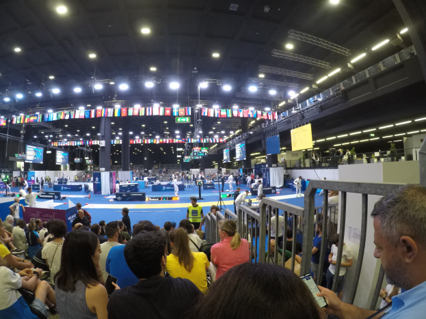 Große Menschenmenge sitzt und steht in einer Indoor-Sportarena mit Bildschirmen, Flaggen und Deckenbeleuchtung.