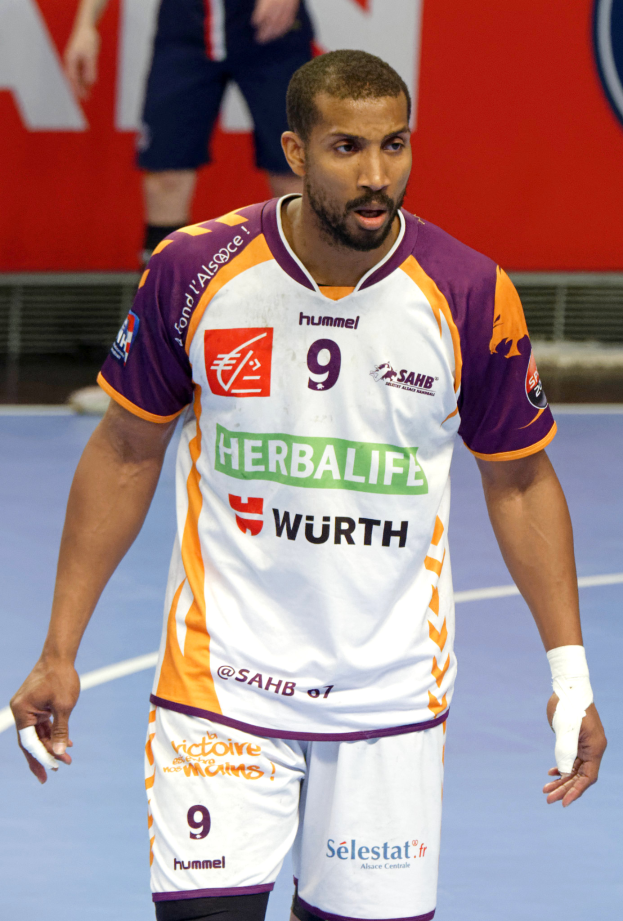 Ein Mann in einem lilafarbenen und weißen Uniform geht auf einem Futsal-Platz, mit zwei Personen im Hintergrund und einer Tafel mit Text darauf, der als Handball-Spieler mit entschlossener Miene erscheint.