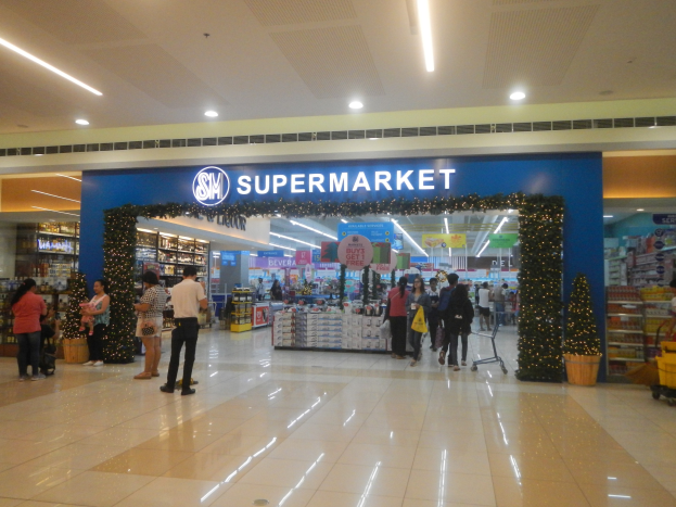 Eine Gruppe von Menschen, die vor einem Supermarkt stehen, einige halten Einkaufstaschen, mit einem Einkaufswagen, einem Topf mit einer Pflanze, Regalen, Schildern und Deckenleuchten im Hintergrund.