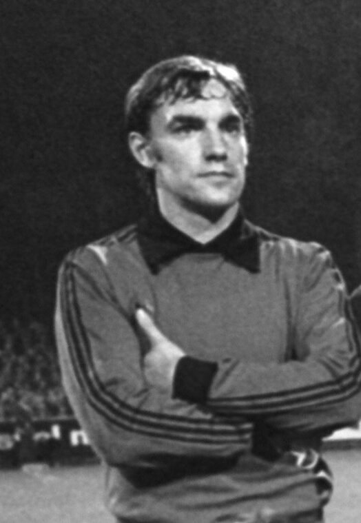 Schwarzes und weißes Foto eines Mannes auf einem Fußballfeld mit verschränkten Armen, umgeben von sitzenden Menschen im Hintergrund.