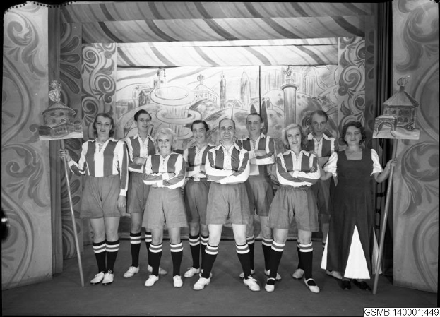 Eine Gruppe von lachenden Frauen in einem Fußballteam der 1940er Jahre posiert auf einer Bühne, jede hält einen Stock in der Hand, mit einer bemalten Wand im Hintergrund.