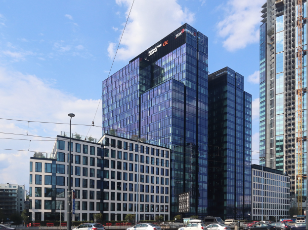 Außenansicht des Deutschen Bank-Hauptsitzes in Frankfurt, Deutschland, mit einem großen mehrfenstrigen Gebäude, umgeben von städtischer Infrastruktur wie Straßenlaternen, Fahrzeugen, Bäumen und einem bewölkten Himmel.
