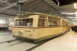 Ein gelb-cremefarbener Zug auf Eisenbahnschienen innerhalb eines Geb├Ąudes, mit einigen Menschen rechts daneben, einer Wand, einem Fenster und Deckenlampen im Hintergrund.