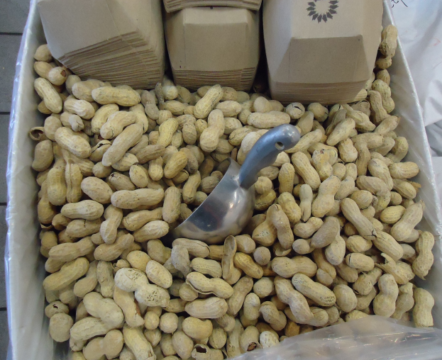 Eine Schachtel Erdnüsse auf einem Tisch mit einer Plastikabdeckung und einem Löffel daneben, umgeben von anderen verstreuten Erdnussboxen.