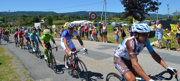Eine Gruppe von Radfahrern fährt auf einer Straße mit Zuschauern an den Seiten, einige tragen Helme und Schutzbrillen und andere halten Fahnen, vor einem Hintergrund aus Bäumen, Schildern, Fahrzeugen, Zelten, Gebäuden, Hügeln und einem klaren blauen Himmel.