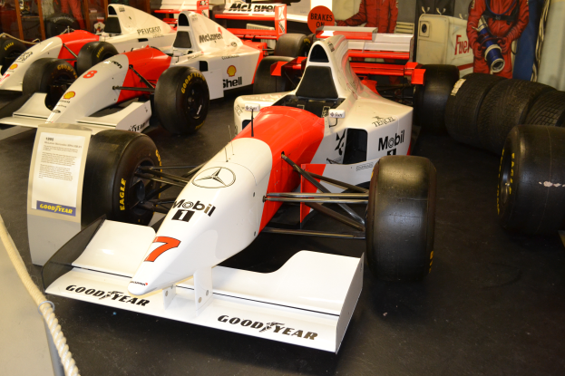 Eine Gruppe von Rennwagen in einem Museum ausgestellt, umgeben von Menschen, ein Schild, ein Seil und eine Wand im Hintergrund.