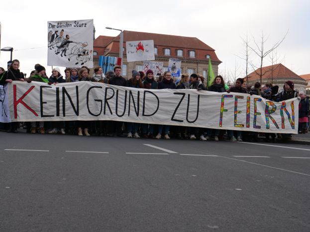 Protestierende mit Transparent 'Kein Grund zur Feier' gegen deutsche Sparmaßnahmen auf einer Stadtstraße mit Gebäuden, Bäumen und einem klaren Himmel.