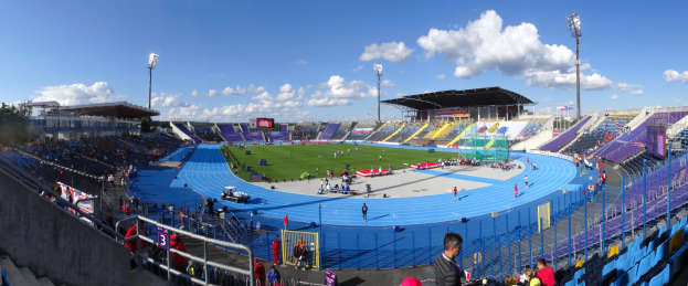 Großes Stadion mit Zuschauern bei einem Leichtathletik-Event, umgeben von einem Zaun, Laternenmasten, Schuppen, Bäumen und Gebäuden unter einem bewölkten Himmel.